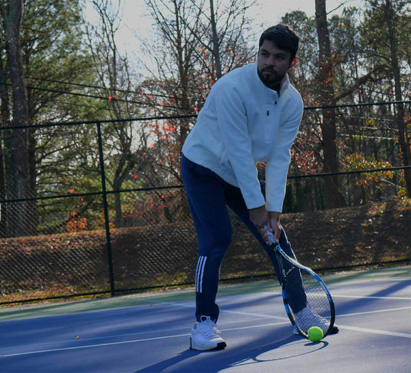 Model bending forward with both hands on a tennis racket, the racket resting on the court surface.